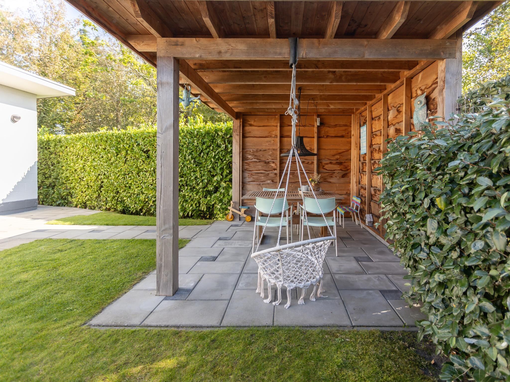 A wooden pavilion with a hanging chair, a table with chairs, and a hedge.