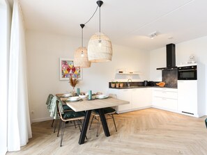 A modern kitchen with white cabinetry, a black stove, and a dining area with a wooden table and chairs. - Modern apartment with balcony by the harbor (Kamperland)