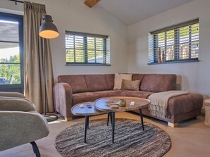 Un salon avec un canapé marron, une table basse ronde, une lampe et deux fenêtres équipées de stores.