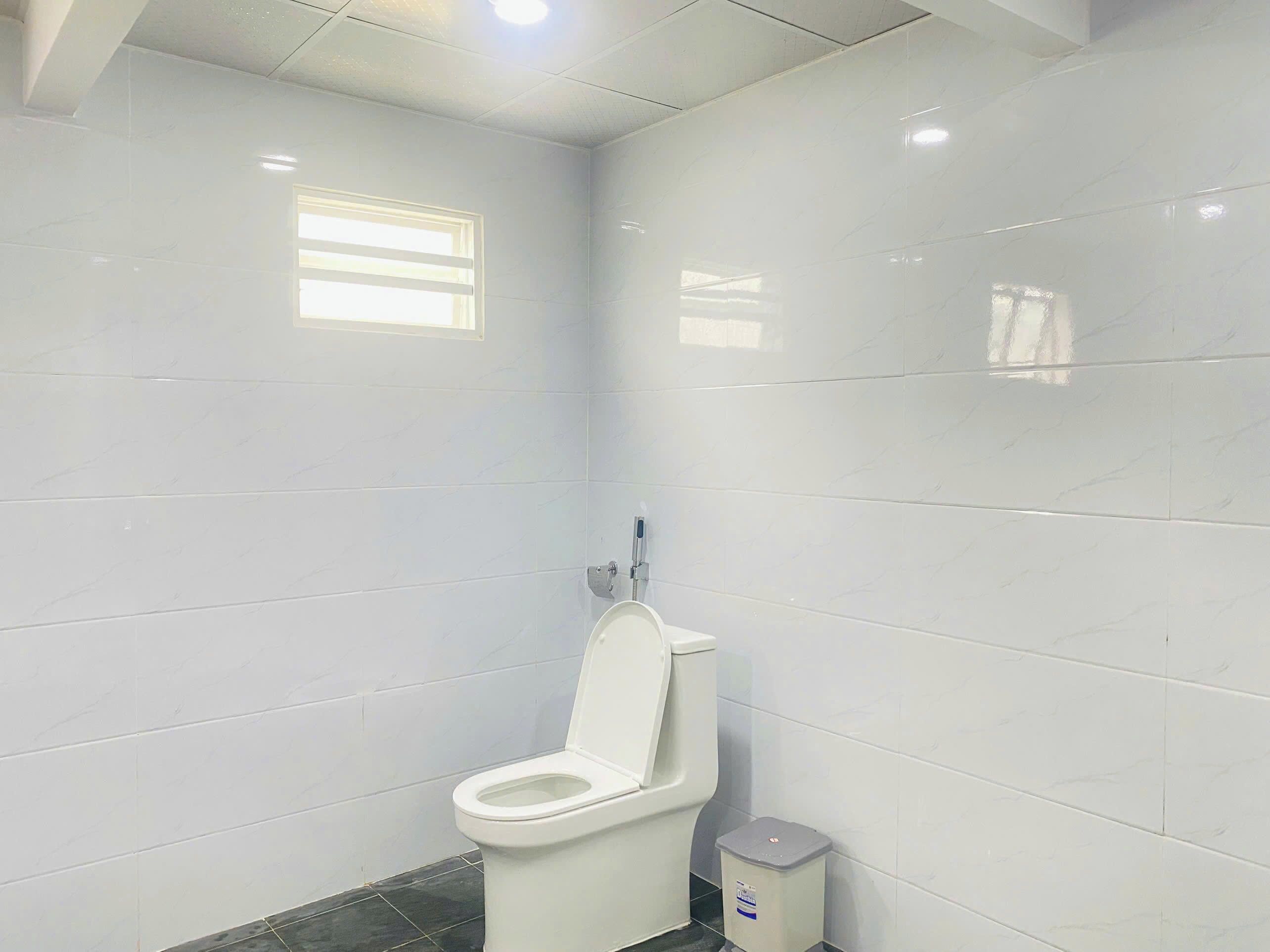 A bathroom with white tiled walls, a white toilet, a trash bin, and a window.