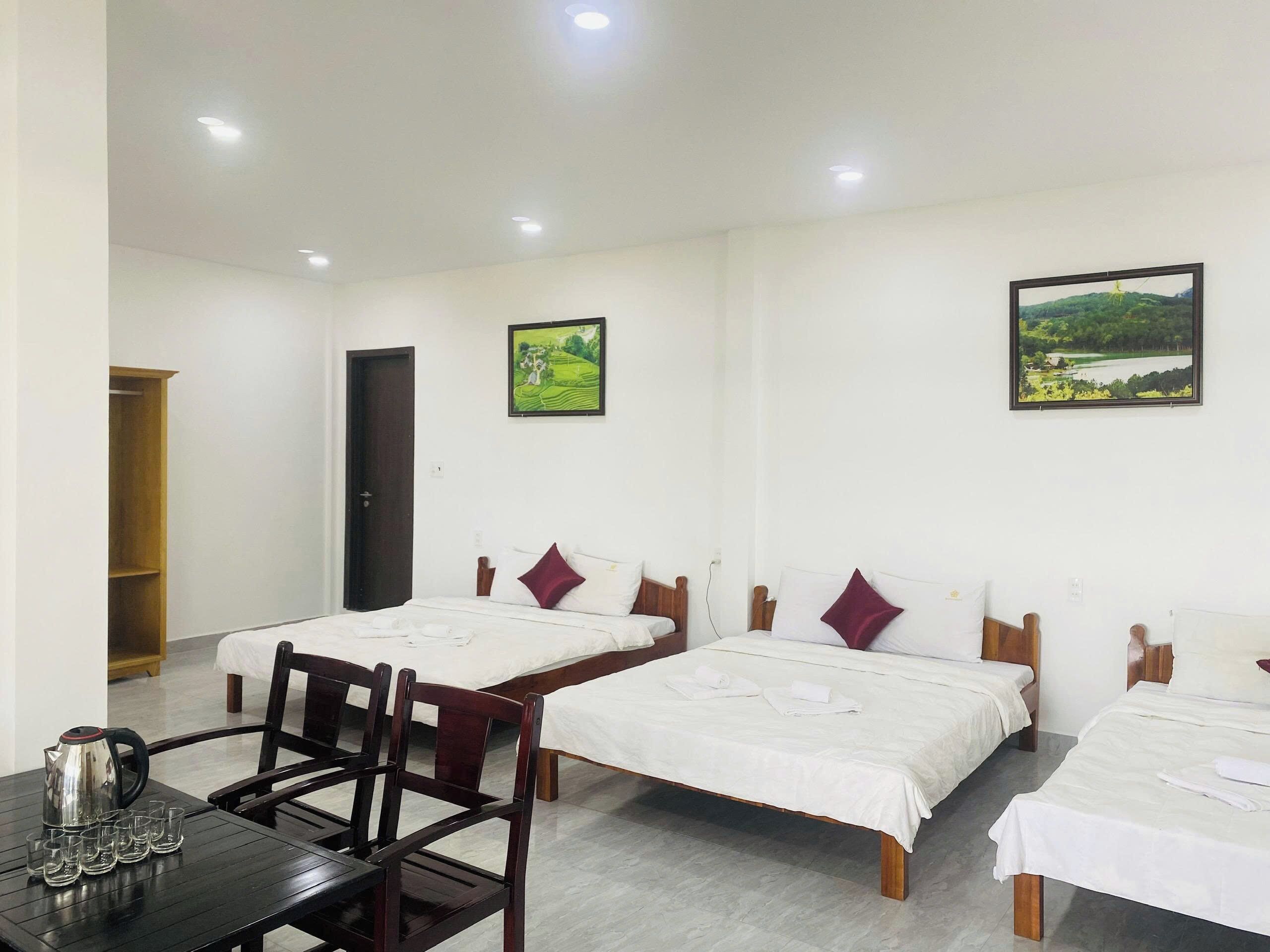 A hotel room with two beds, a wooden table with a kettle and glasses, and two framed landscape pictures on the wall.