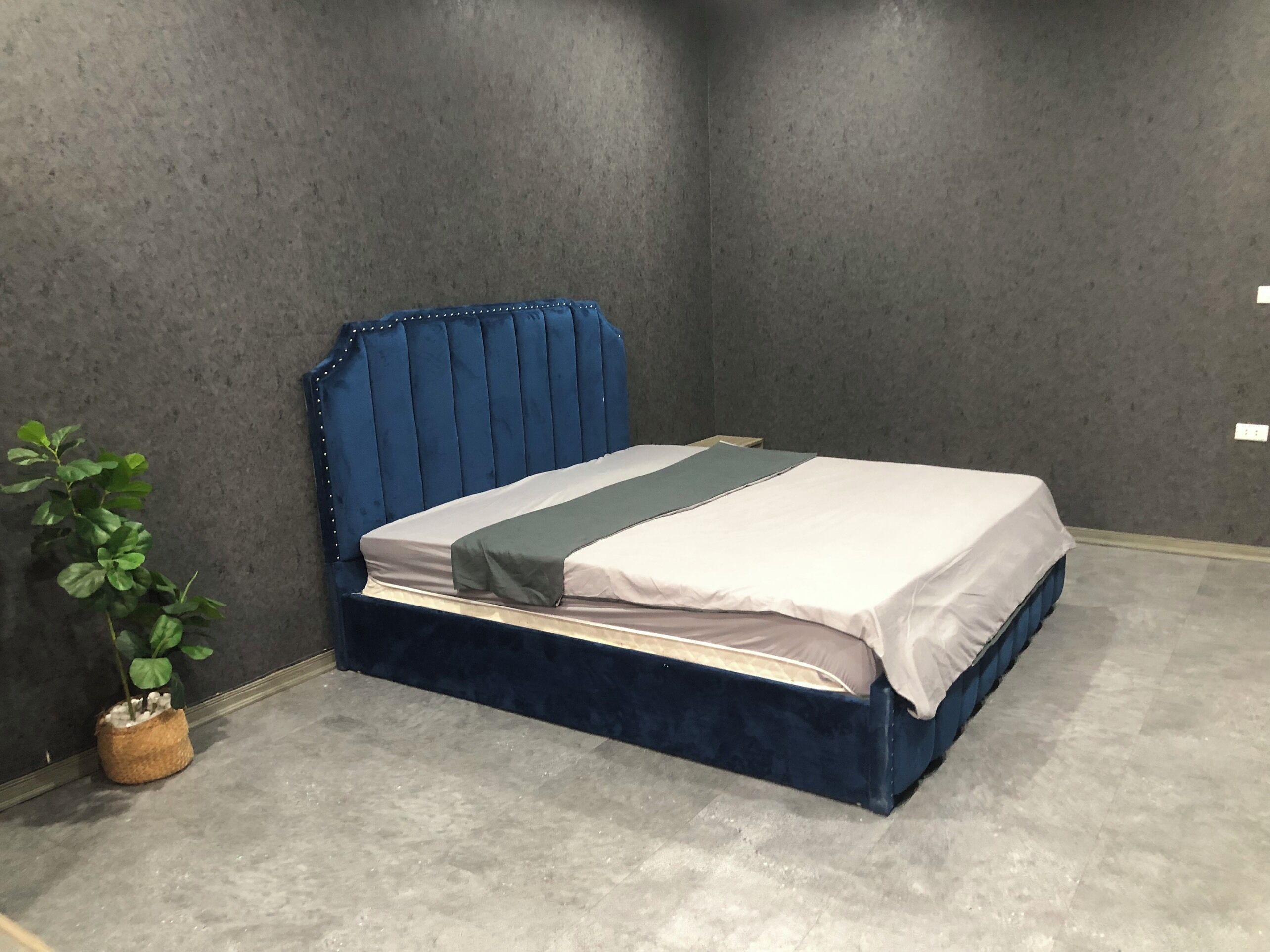 A bed with a blue upholstered headboard in a room with grey walls and a concrete floor.