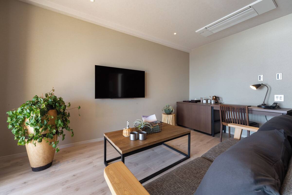 Ein modernes Wohnzimmer mit Flachbildfernseher, Couchtisch aus Holz, einer Bank, einer großen Topfpflanze und einer kleinen Küchenzeile mit Schreibtisch und Stuhl.