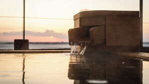 Estructura de madera con una fuente de agua, reflejada en una superficie pulida, y una vista del atardecer al fondo.
