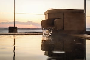Estructura de madera con una fuente de agua, reflejada en una superficie pulida, y una vista del atardecer al fondo.