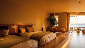 Habitación de hotel con dos camas, una zona de estar, un ventanal con vistas y una planta en maceta.