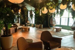 A spacious indoor area with a variety of seating options, including wicker chairs and a hanging chair, surrounded by large potted plants and hanging lanterns. - Skyline Retreat 1BR+Den | Walkable Downtown + Pool (Miami)
