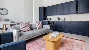 Un salon moderne avec un canapé gris, une table basse en bois et un coin cuisine doté de meubles sombres et d’appareils électroménagers modernes.