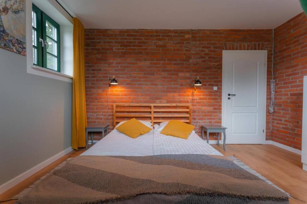 Une pièce avec un mur en briques, un lit avec une literie blanche et des oreillers jaunes, deux tables de chevet, une fenêtre avec des rideaux jaunes et une porte blanche.