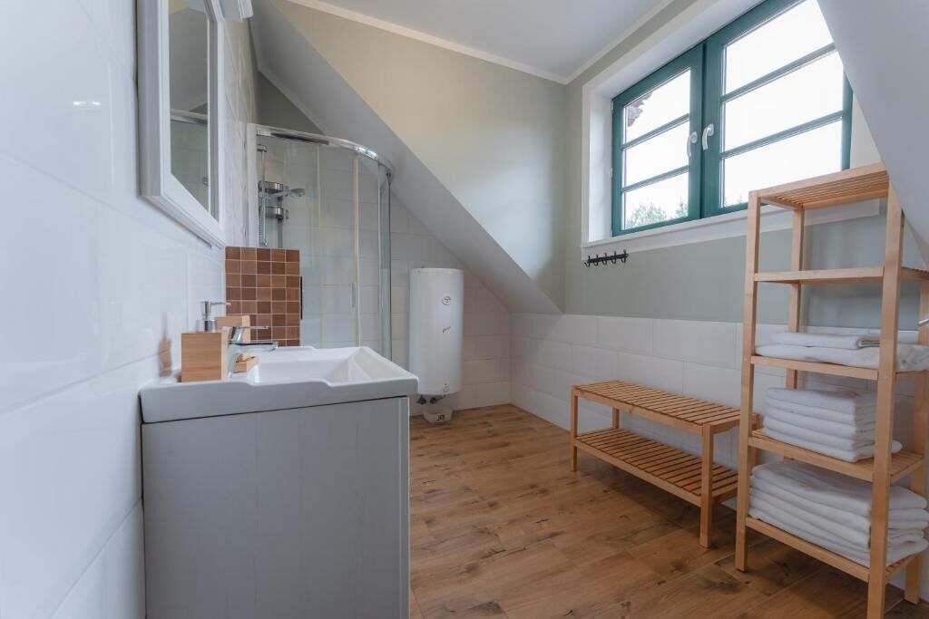 A bathroom with a shower, a sink, a toilet, a wooden shelf with towels, and a window with a view.