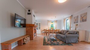 A living room with a fireplace, a sofa, a TV mounted on the wall, and a dining area with chairs and a table. - Mazurskie Domy Nad Kisajnem (Giżycko)
