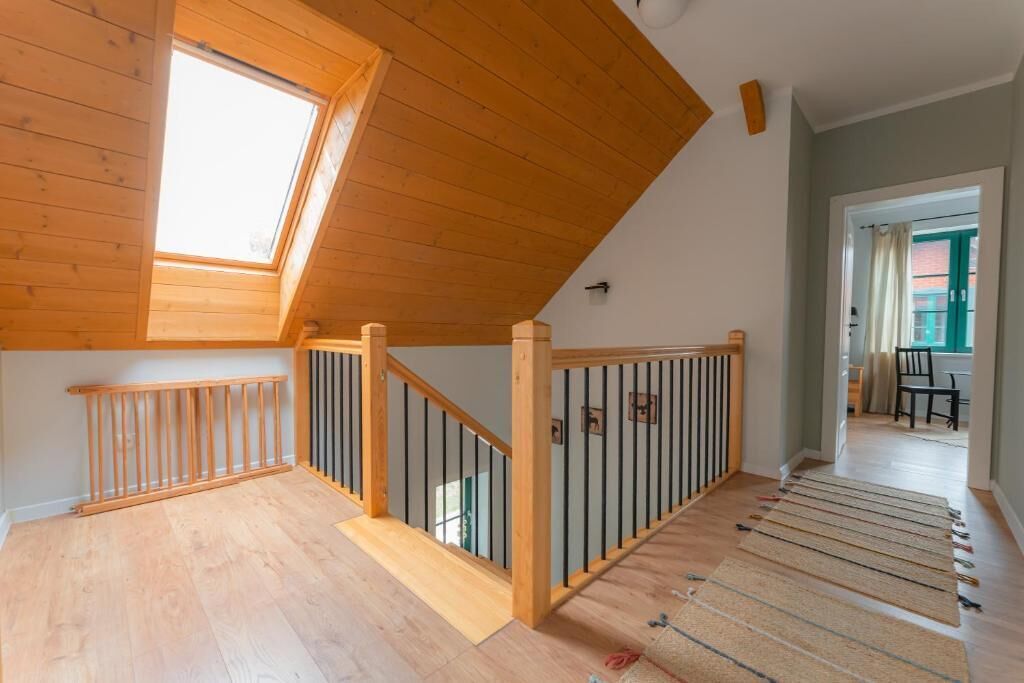 Une pièce avec un plafond en pente, un plancher en bois, un escalier avec une rampe et une vue sur l’extérieur par une porte ouverte.