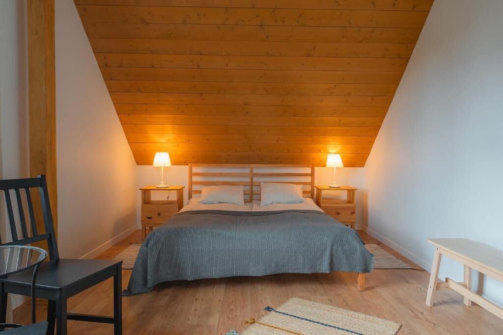 A bedroom with a sloped ceiling, a bed with a gray bedspread, two bedside lamps, a chair, a small table, and a wooden bench.