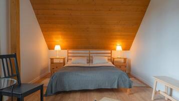 A bedroom with a sloped ceiling, a bed with a gray bedspread, two bedside lamps, a chair, a small table, and a wooden bench.
