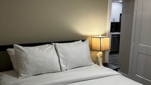 Una habitación de hotel con una cama, una mesita de noche con lámpara y una puerta que da a una cocina americana.
