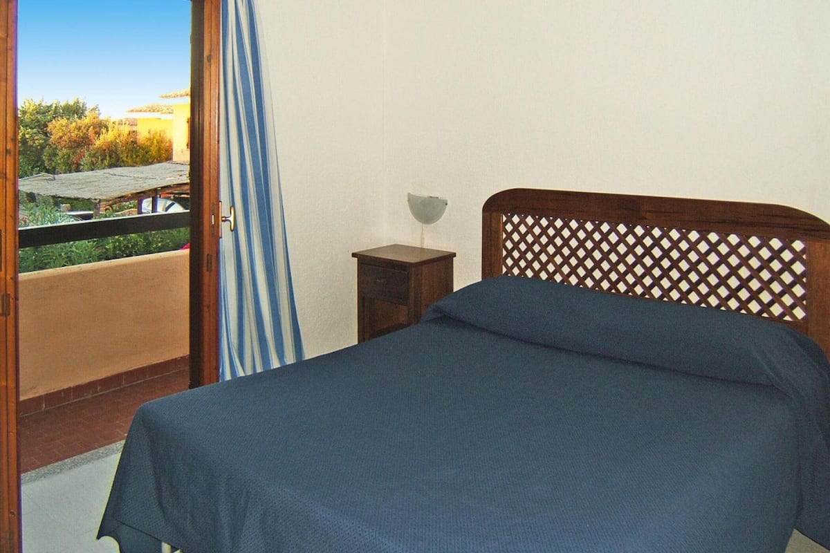 A bedroom with a bed, a nightstand, a window with a view of greenery, and a balcony.