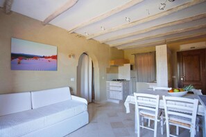A room with a white sofa, a dining table with chairs, a kitchen area, and a painting on the wall. - Holiday Resort Baia de Bahas Residence, Golfo Aranci (Golfo Aranci)