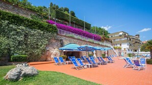 Ein Poolbereich mit Liegestühlen und Sonnenschirmen, eine Steinwand mit Bepflanzung sowie ein Gebäude im Hintergrund.