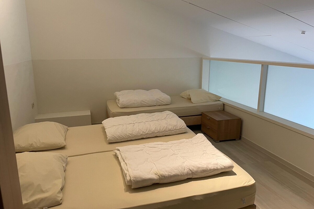 Ein modernes, minimalistisches Schlafzimmer mit zwei Betten, einem Nachttisch und einem Fenster, das natürliches Licht hereinlässt.