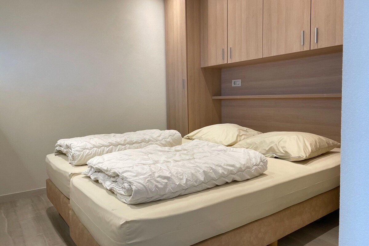 Ein Einzelbett mit beiger Matratze und weißer Bettwäsche, ein Holz-Kopfteil und ein kleines Regal darüber.