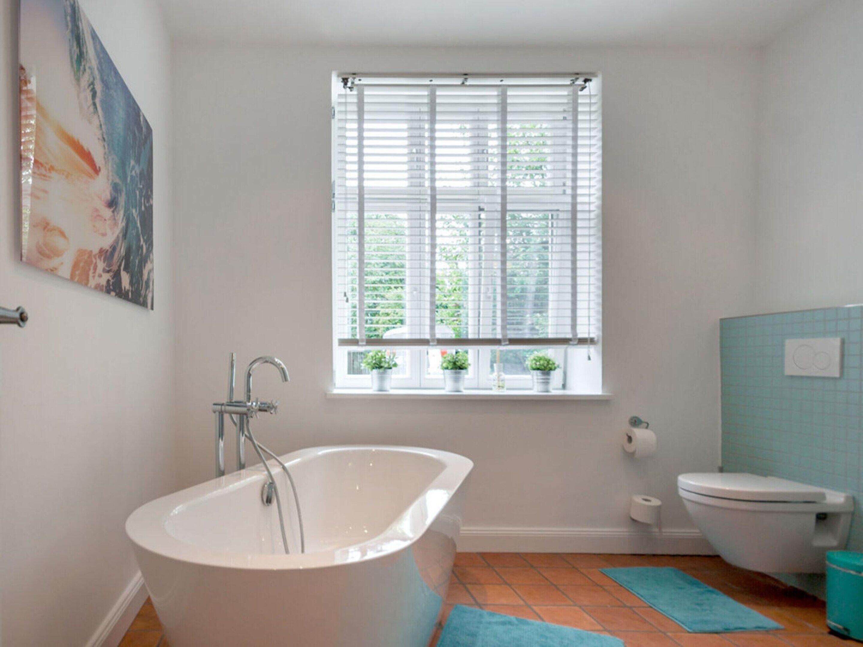 Ein Badezimmer mit freistehender Badewanne, Toilette, einem Fenster mit Jalousien und einem kleinen Topfpflanzen auf dem Fensterbrett.