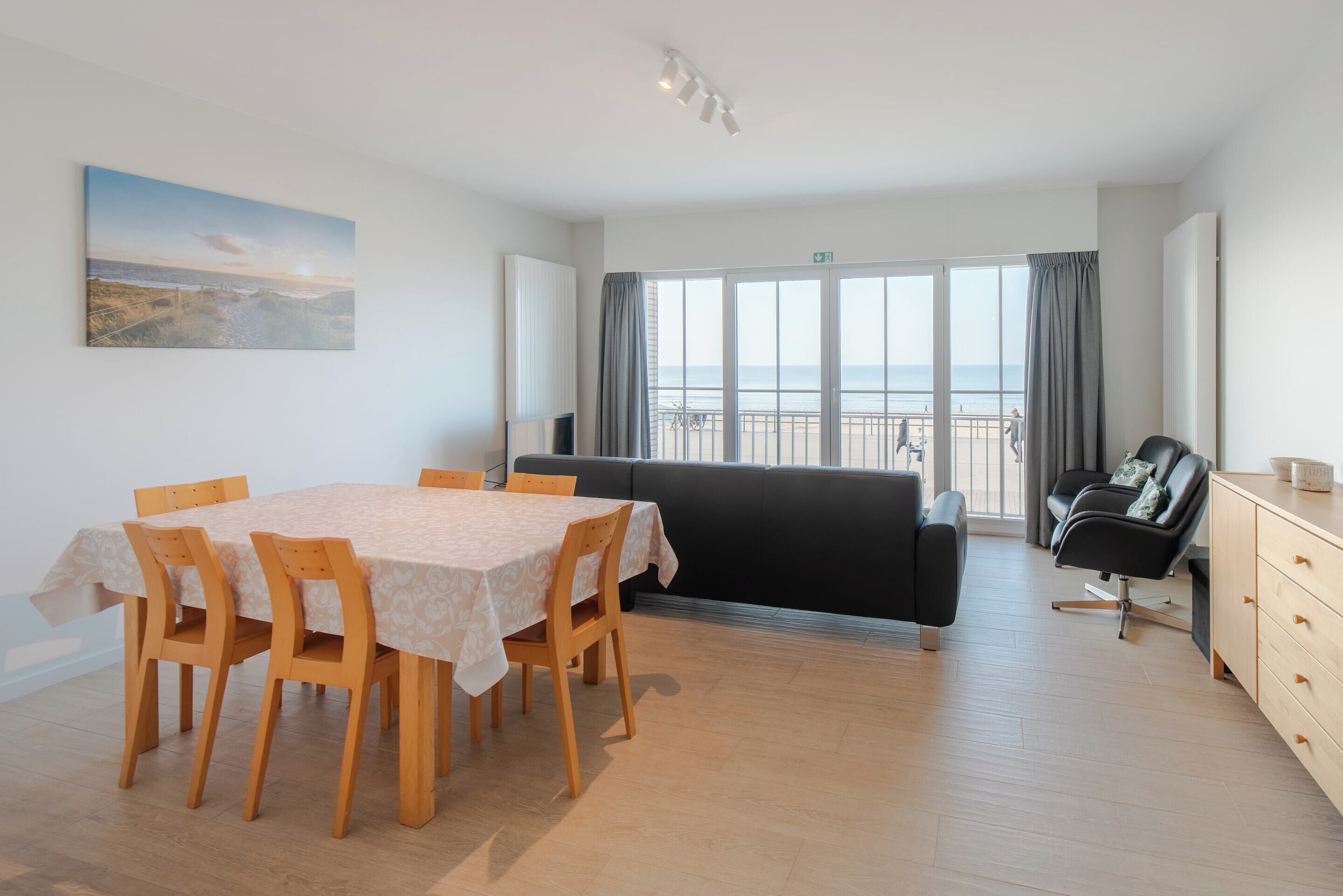 Un salon moderne comprenant un coin repas, un canapé noir, un meuble en bois et une grande fenêtre donnant sur la plage.