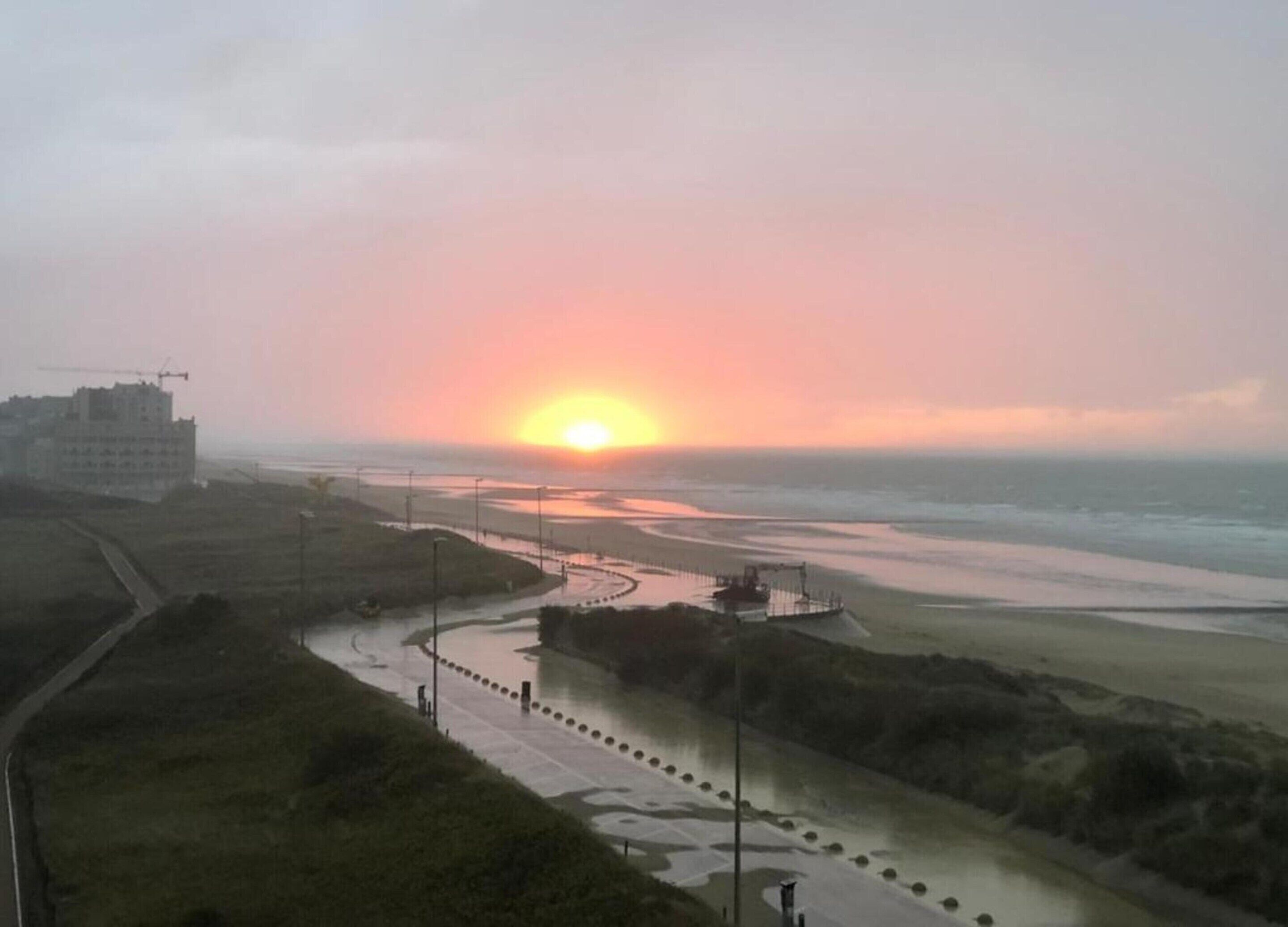 Une vue sur le coucher de soleil sur la côte, avec une route menant à la plage, un chantier de construction et un bâtiment au loin.