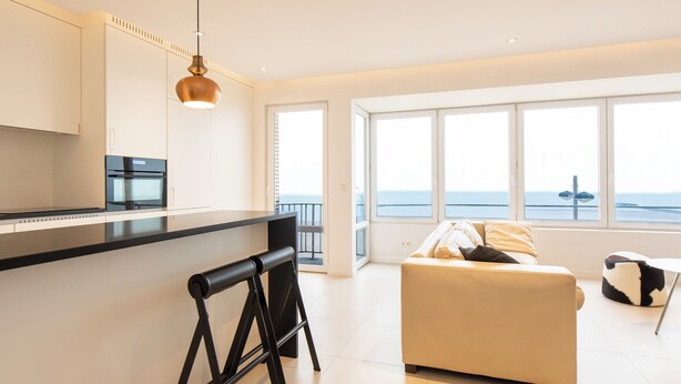 Une cuisine moderne avec un plan de travail noir, un luminaire suspendu et une vue sur la mer grâce à de grandes fenêtres.