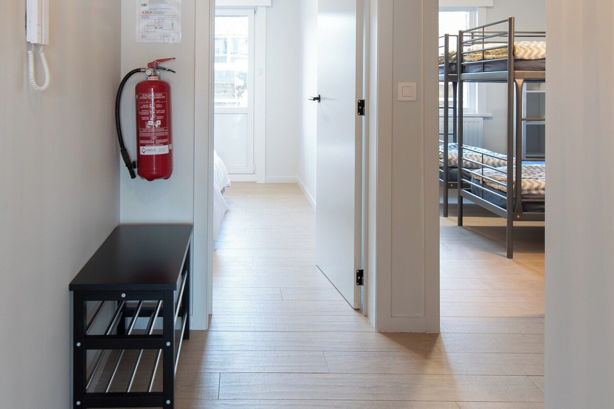 A hallway with a fire extinguisher, a bench, and bunk beds in the background.