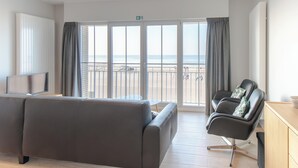Une chambre d’hôtel moderne, dotée d’un canapé, d’un fauteuil, d’une télévision et offrant une vue sur la plage grâce à de grandes fenêtres.