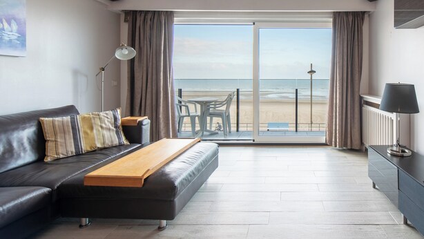 A modern living room with a sofa, a wooden table, a lamp, and a view of the beach through a glass door. - Apartment in Middelkerke With Sea View (Middelkerke)
