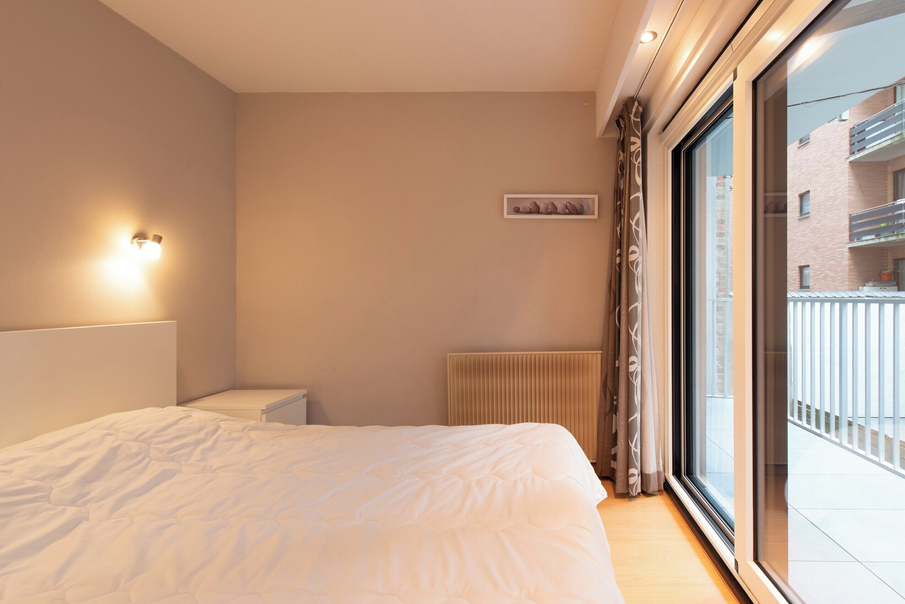 A bedroom with a bed, a radiator, a wall-mounted light, and a window with a view of a building.