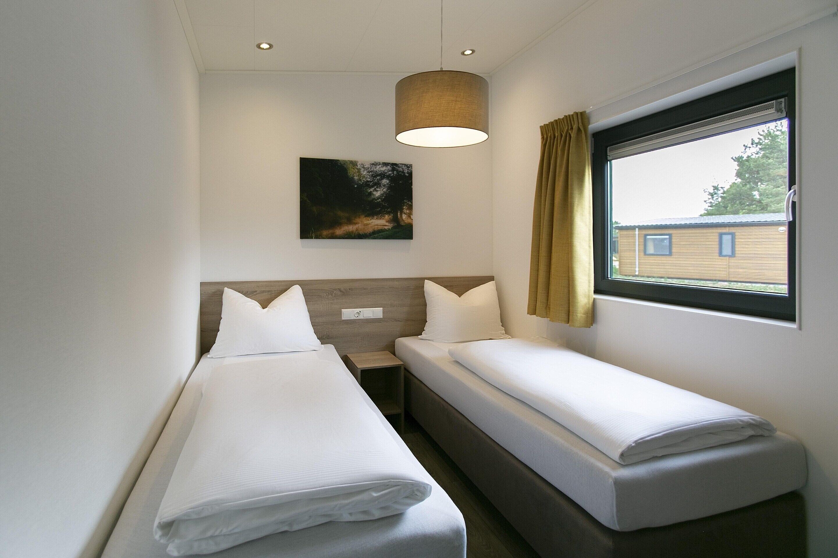 Une chambre d’hôtel avec deux lits simples, une table de chevet, une lampe fixée au mur, une fenêtre avec des rideaux et un tableau accroché au mur.
