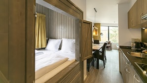 A bedroom with a wooden headboard, a bed with white linens, a dining area with a table and chairs, and a kitchen with a sink and stove. - Mobile Home im Tropical Islands Resort (Halbe)