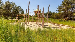 A playground with a climbing structure and swings, surrounded by tall trees and grassy areas. - Mobile Home im Tropical Islands Resort (Halbe)