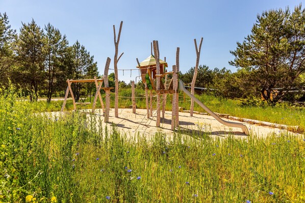 Une aire de jeux avec une structure d’escalade et des balançoires, entourée de grands arbres et de zones herbeuses.
