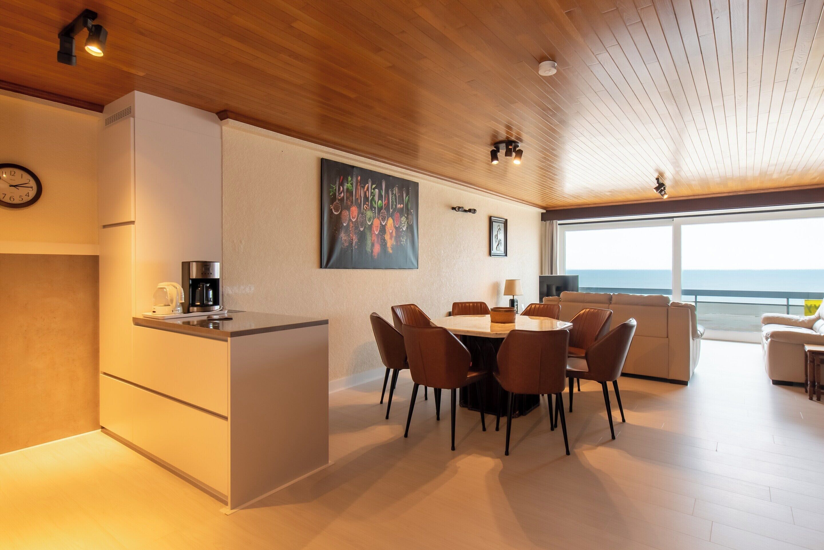 A modern kitchen with a dining area, a large painting, and a view of the sea.