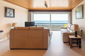 A modern living room with a sofa, a smaller couch, a TV, and a table with a lamp. A large window offers a view of the ocean. - Apartment in Middelkerke, Beach Views (Middelkerke)