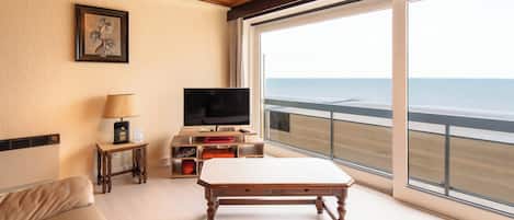 A room with a wooden ceiling, a television, a table, a sofa, and a large window with a view of the ocean.