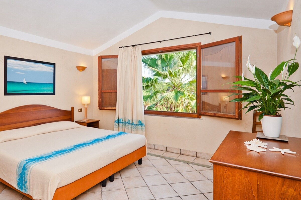 A hotel room with a bed, bedside lamps, a window with curtains, a potted plant, and a wall-mounted TV.