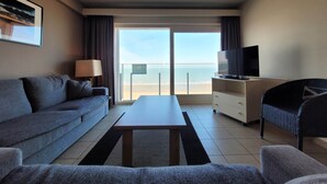 A hotel room with a sofa, a coffee table, a TV on a stand, a chair, and a balcony with a view of the beach. - Steps From the Quiet Seaside Path (Middelkerke)