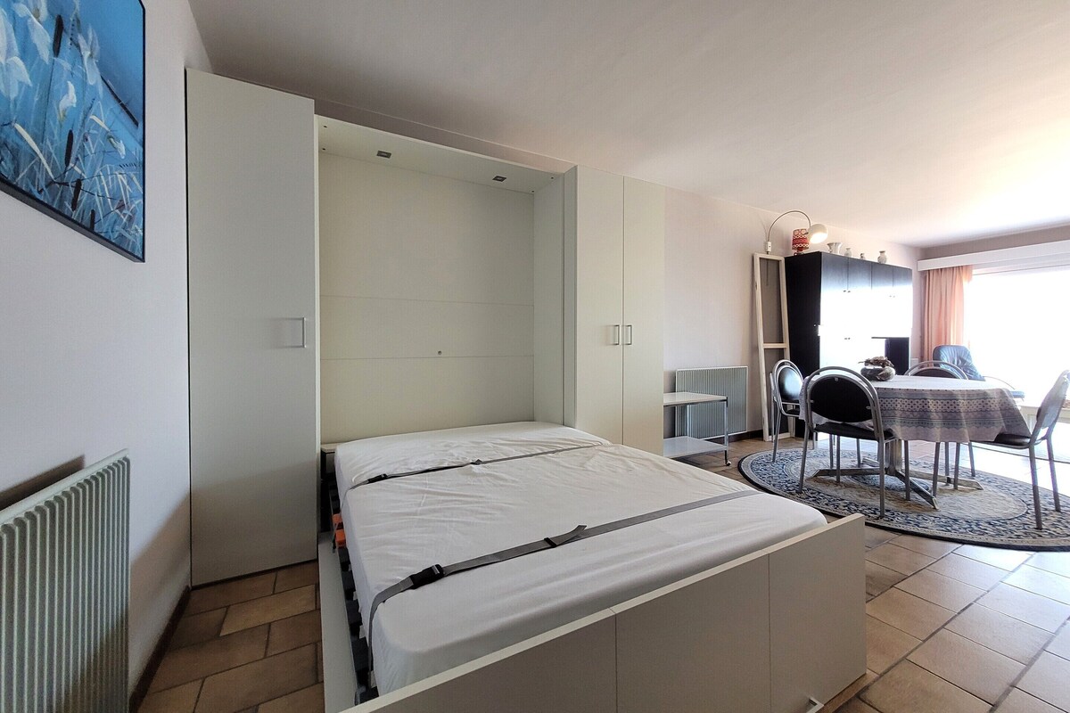A compact room with a bed, a desk, and a chair. There is a radiator under the bed and a window with curtains.