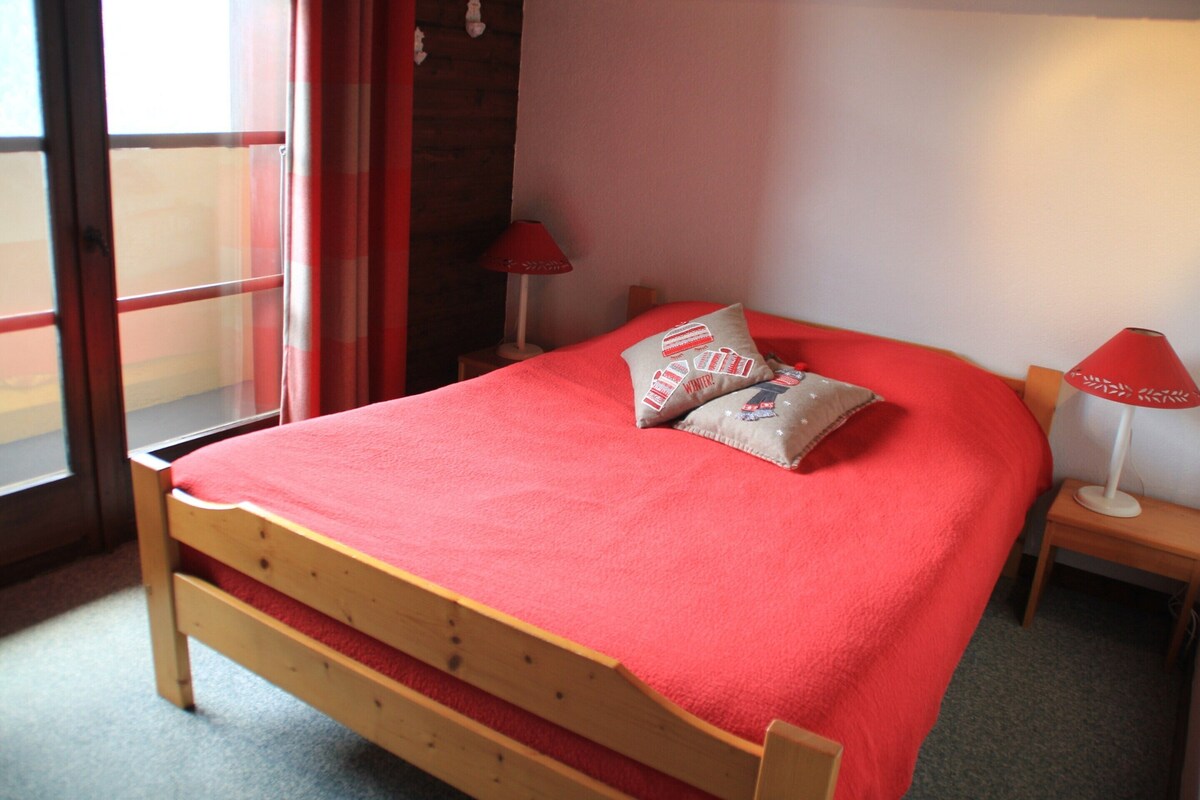 A bedroom with a wooden bed, red bedding, two pillows, a nightstand with a lamp, and a window with red curtains.