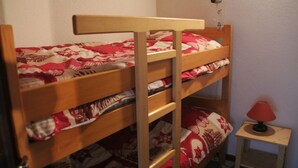A bunk bed with a red and white patterned bedspread, a small bedside table with a lamp, and a wall-mounted heater. - Apartment With Parking for 6 People (Chatel)