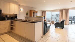 A modern kitchen with white cabinets, a black countertop, and a built-in island. There is a dining area with a gray sofa and a small table. The room has large windows with curtains and a view of buildings. - Apartment in Middelkerke Near Casino (Middelkerke)
