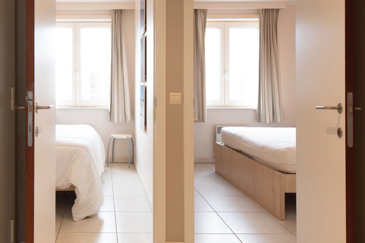 Ein Hotelzimmer mit Bett, einem kleinen Hocker und einem Fenster mit Vorhängen.