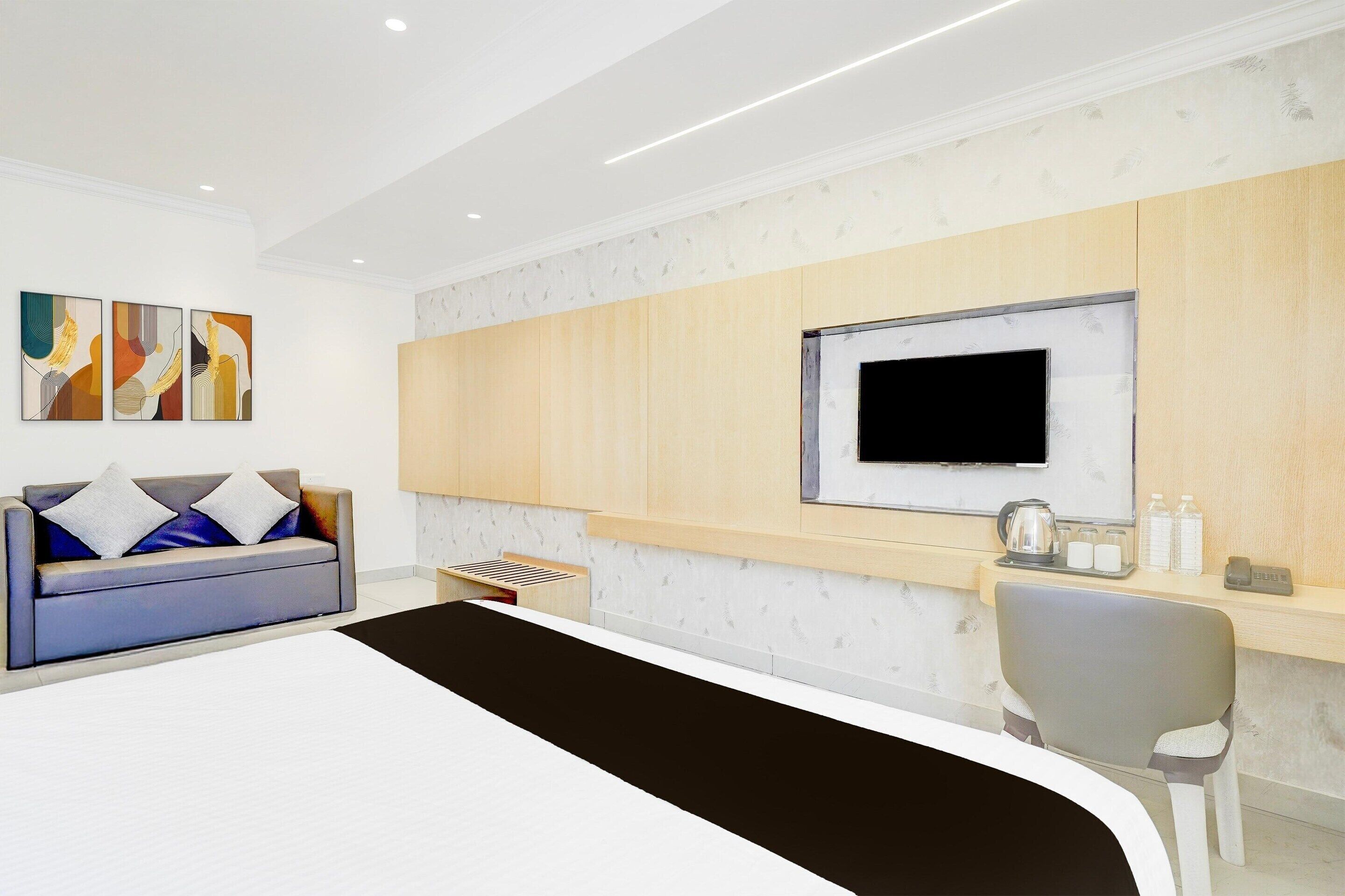 A modern hotel room with a sofa, a flat-screen TV, a desk with a kettle, and a chair.