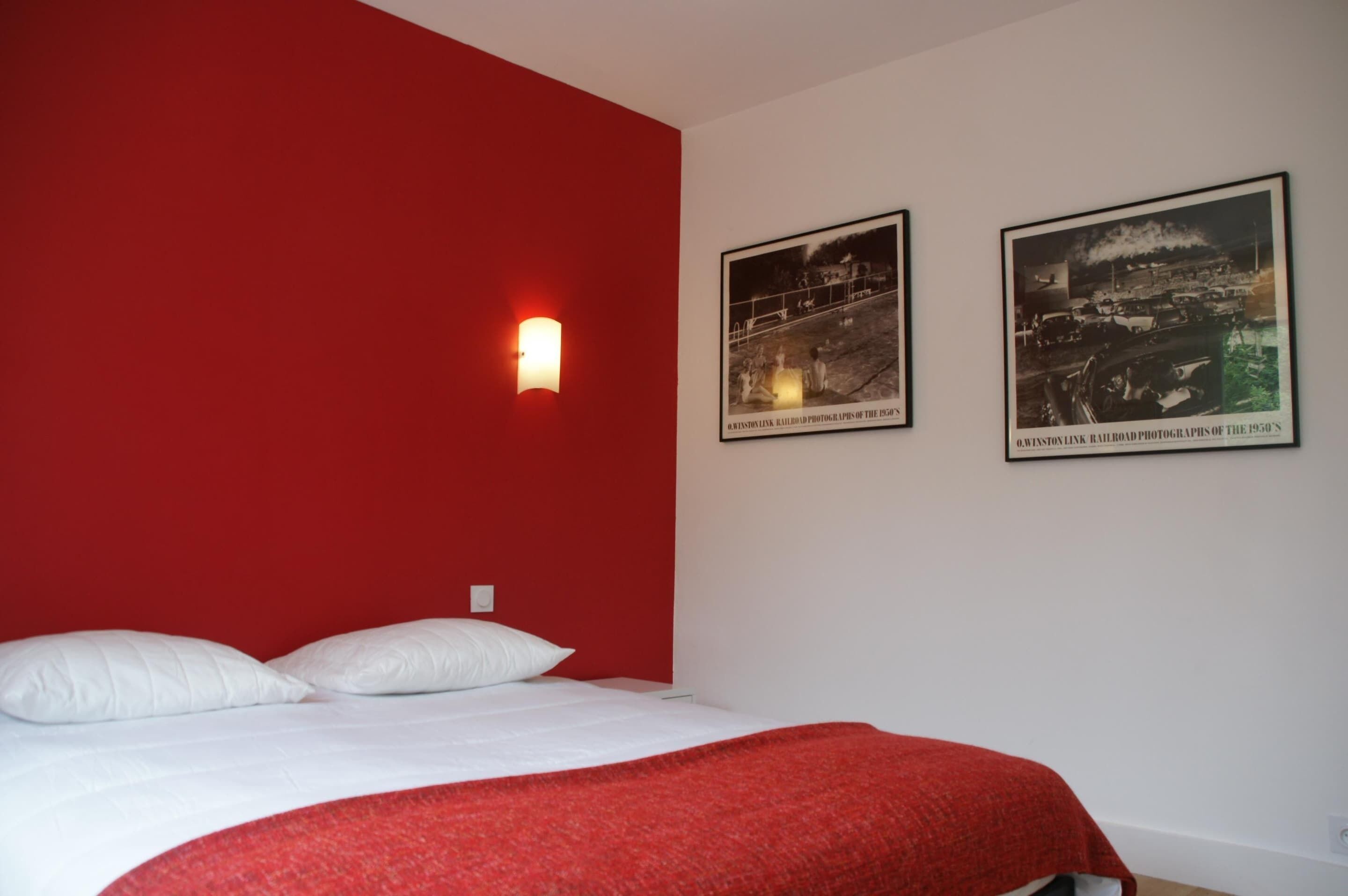 Et hotelværelse med en rød accentvæg, en seng med hvide sengetøj og to sort-hvide fotografier på væggen.