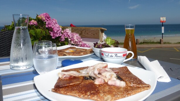 A meal setup with a plate of food, a glass of water, and a bottle of beer on a table with a view of the beach and ocean. - Family Home in Locquirec Bay (Locquirec)