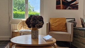 A room with a round table, a chair, a vase with dried flowers, a book, and a window with a view of greenery. - Family Home in Locquirec Bay (Locquirec)